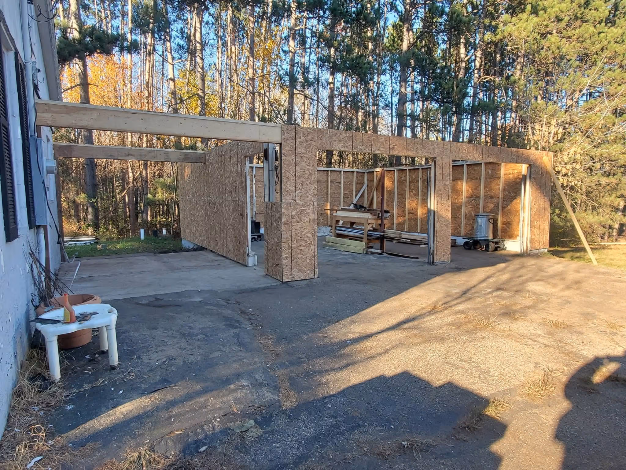 Partially built wooden garage frame attached to a house, with tools and materials inside, set in a wooded area.