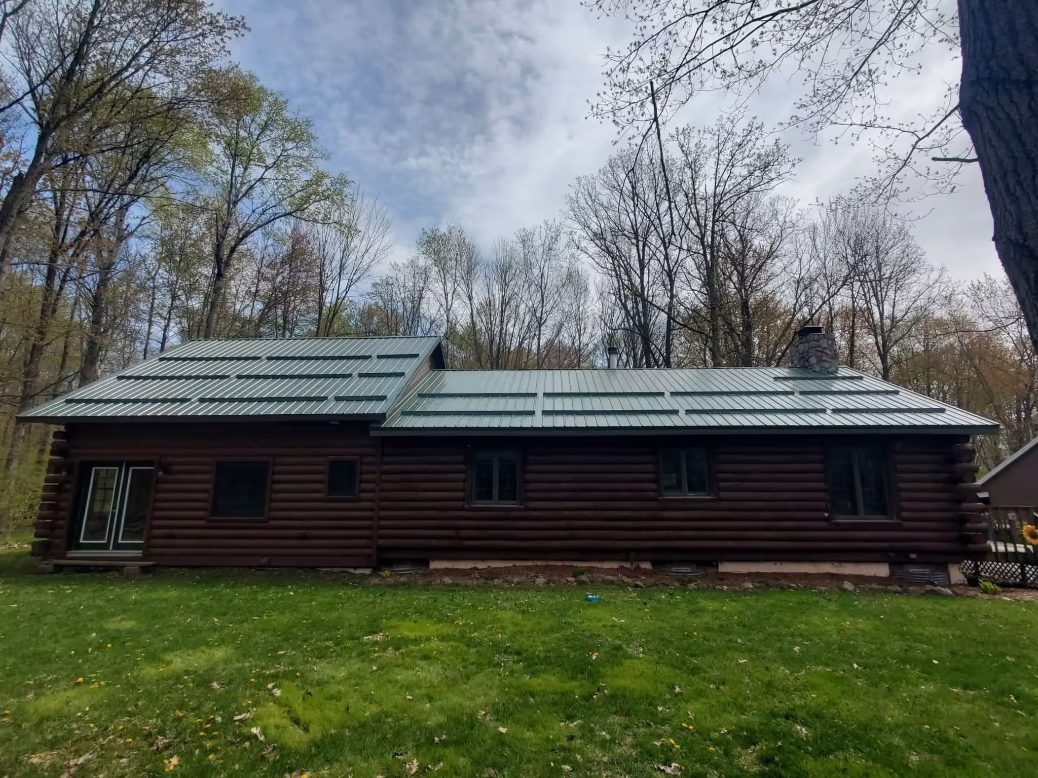 Log cabin with a green metal roof in a grassy yard surrounded by tall trees with sparse leaves.