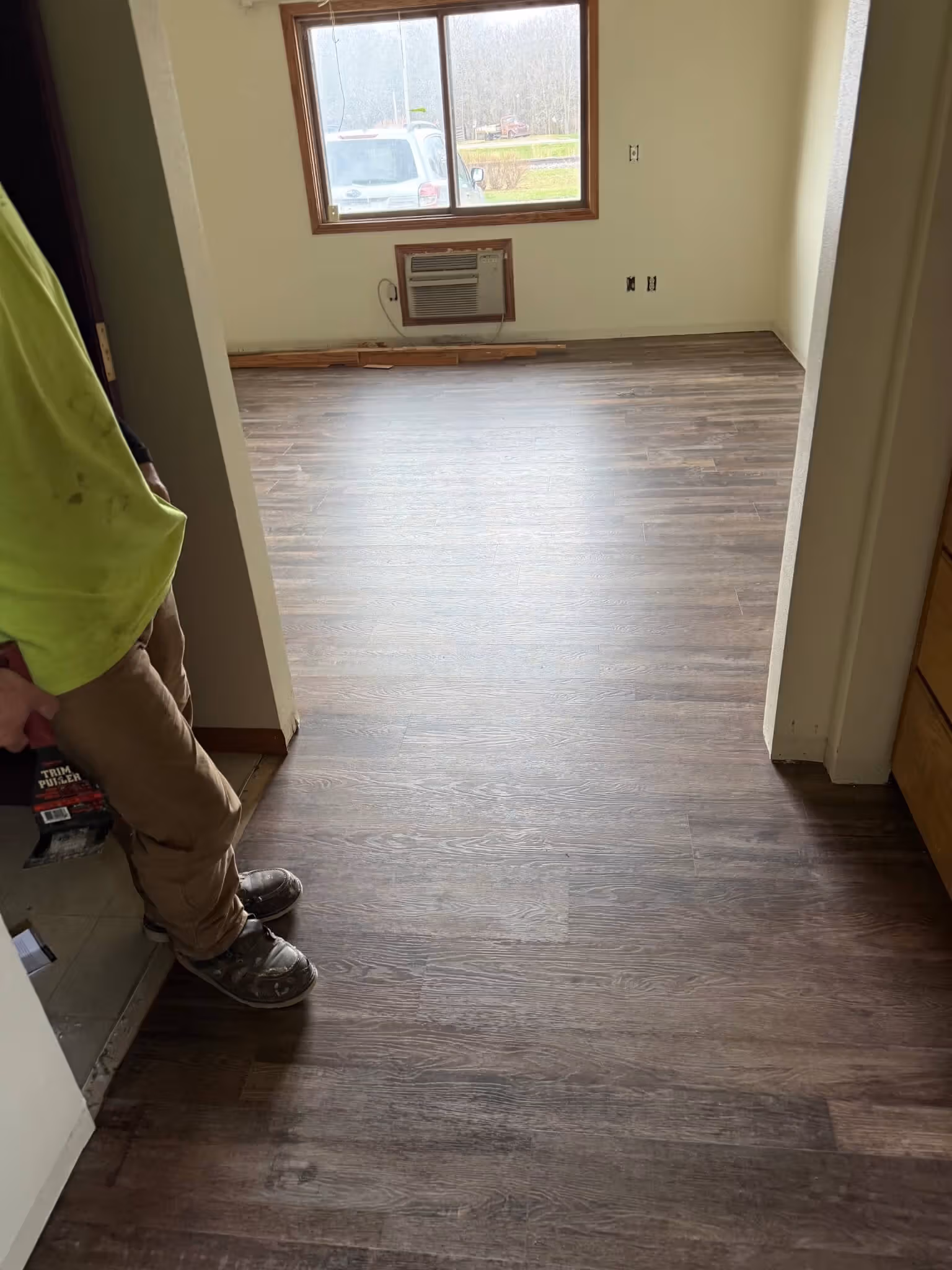 Person standing at the edge of new dark wood laminate flooring in an empty room with a window AC unit.