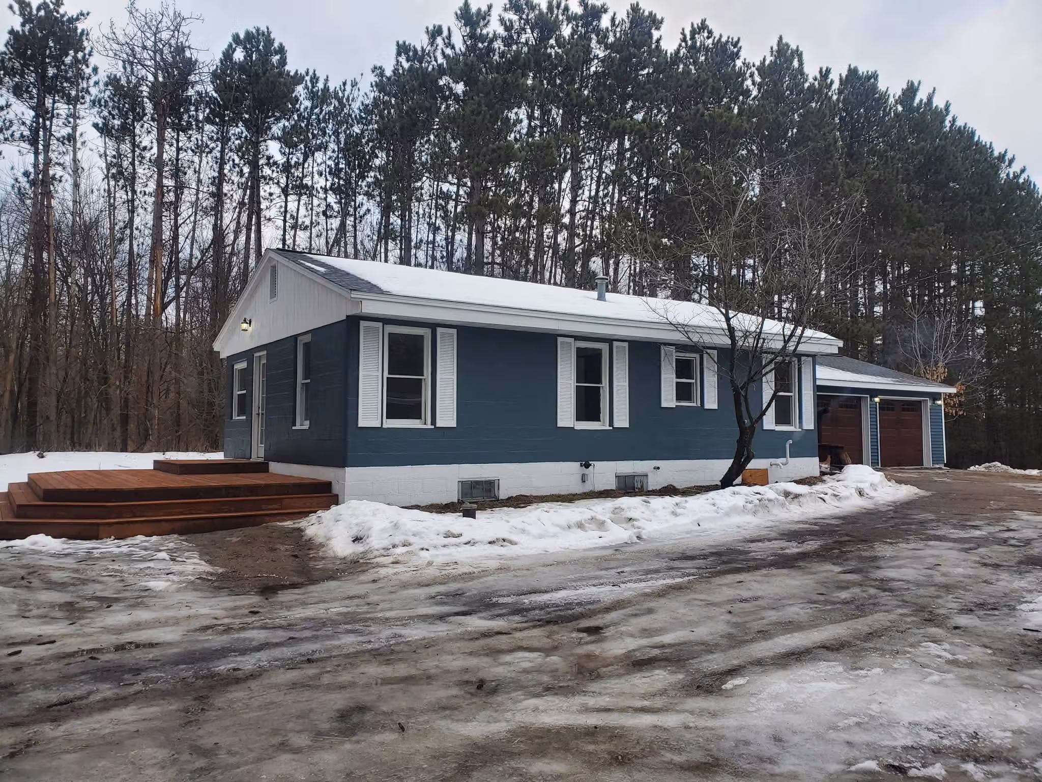 Single-story blue house with white shutters, wooden deck, and garage in a snowy landscape with tall pine trees.