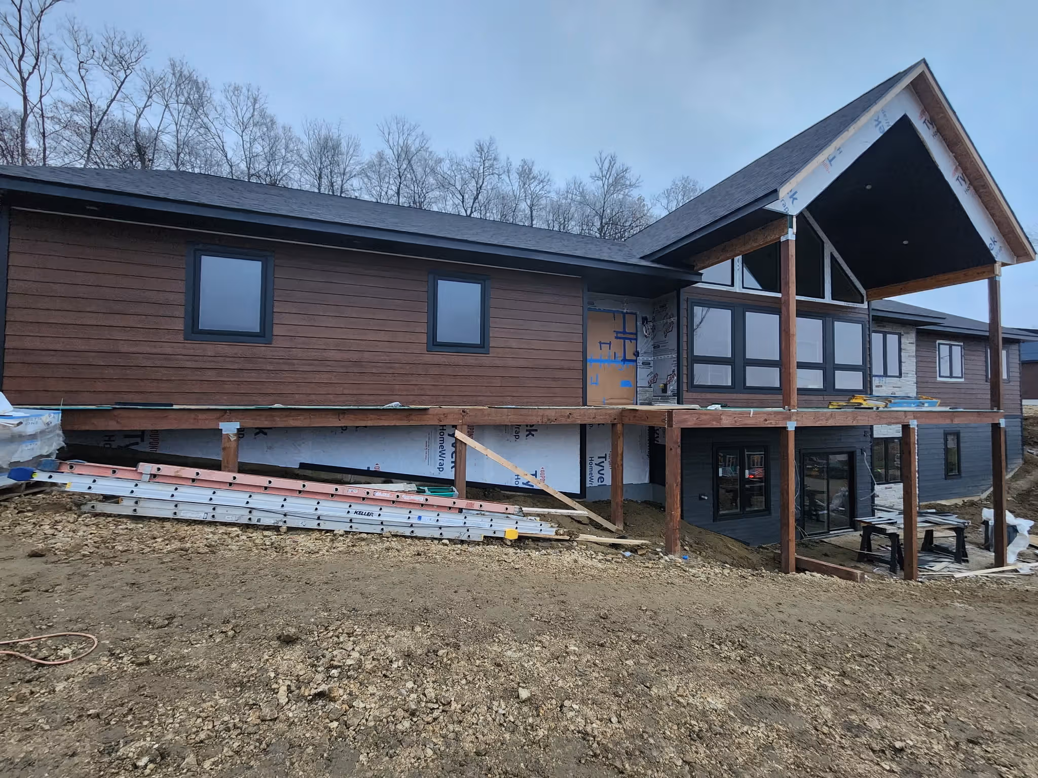 Modern house under construction with wood siding, large windows, and an elevated porch framework.