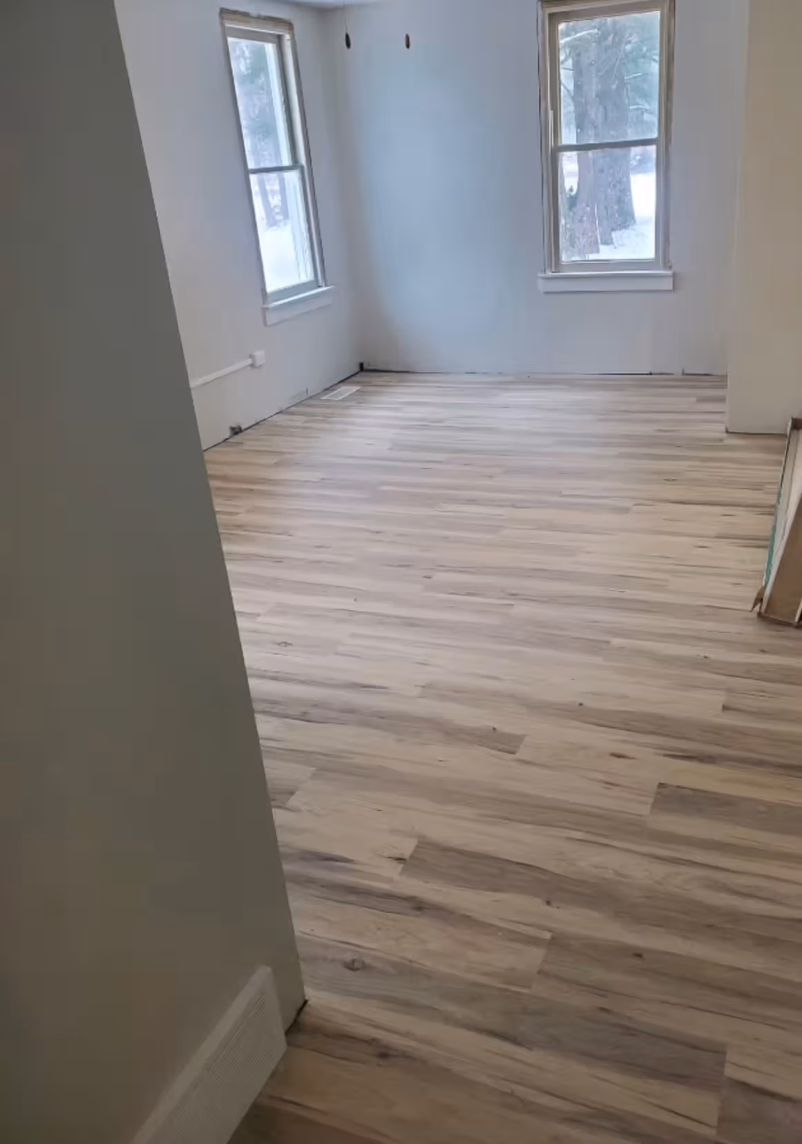 Empty room with light wood laminate flooring and two large windows showing snowy trees outside.
