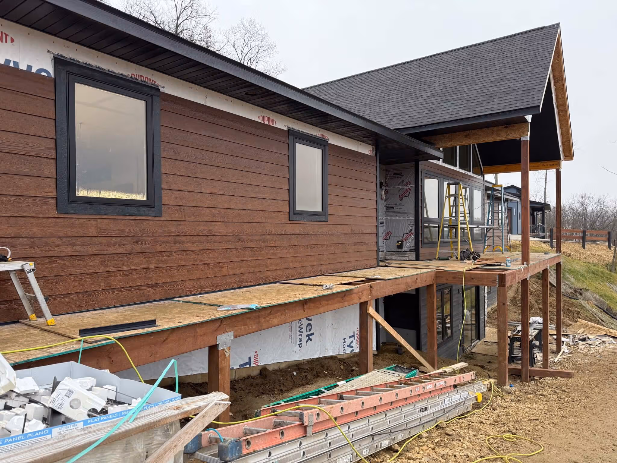 Construction site of a modern house with wooden siding and an incomplete raised deck with ladders and tools.