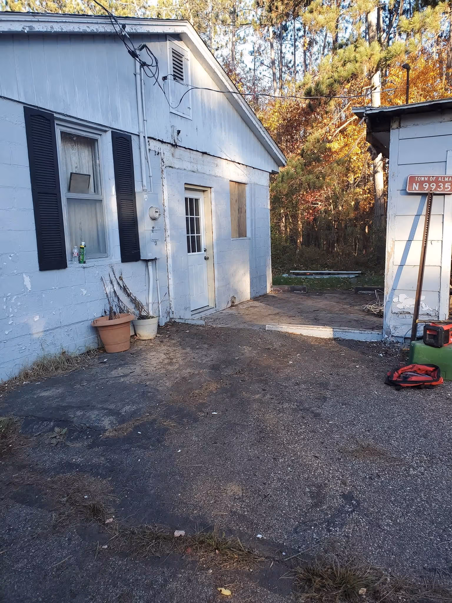 Small white building with black shutters, a white door, potted plants, and a gravel driveway surrounded by trees.