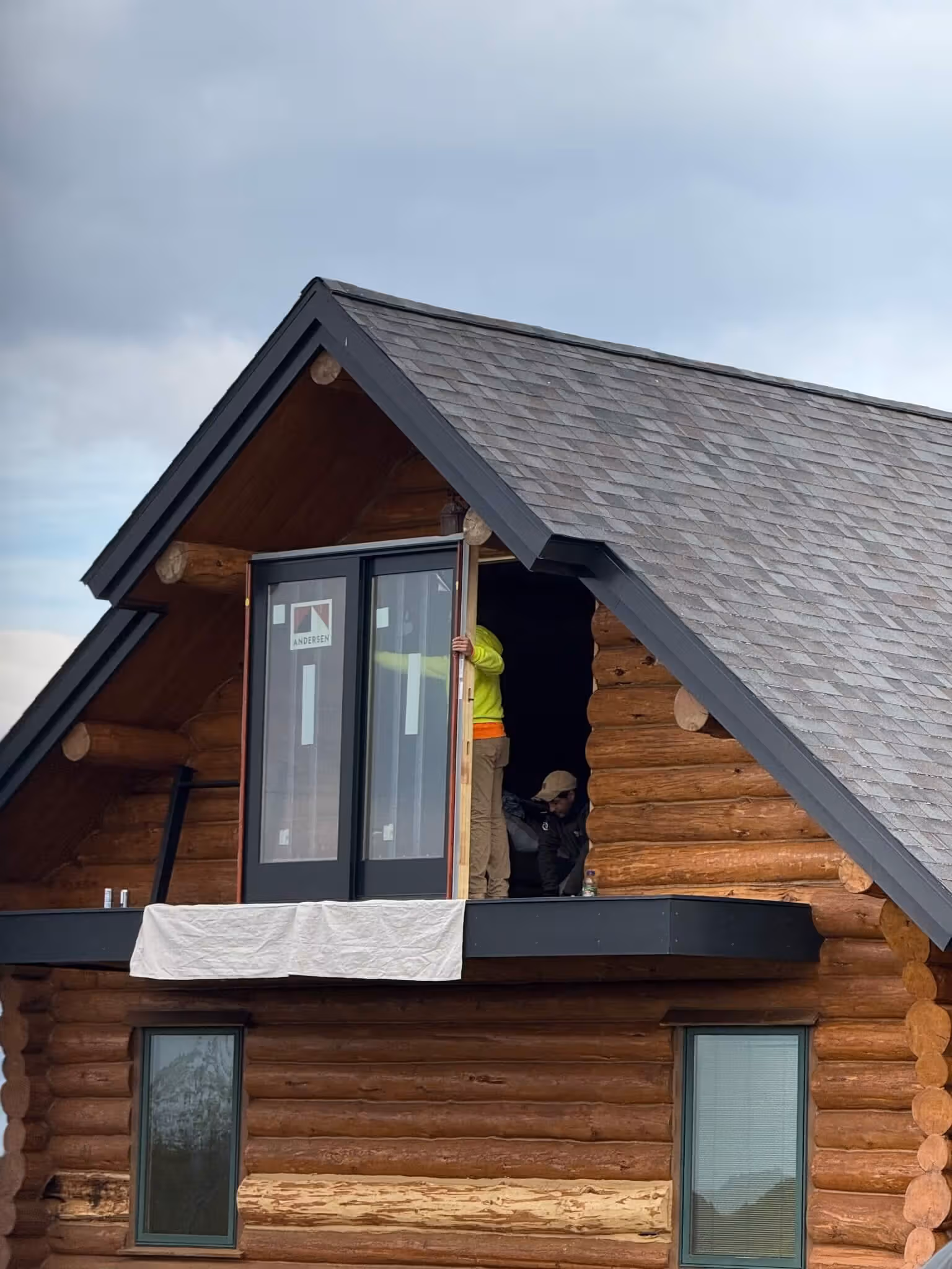 Two workers installing glass doors on the upper balcony of a wooden log cabin under a gray sky.
