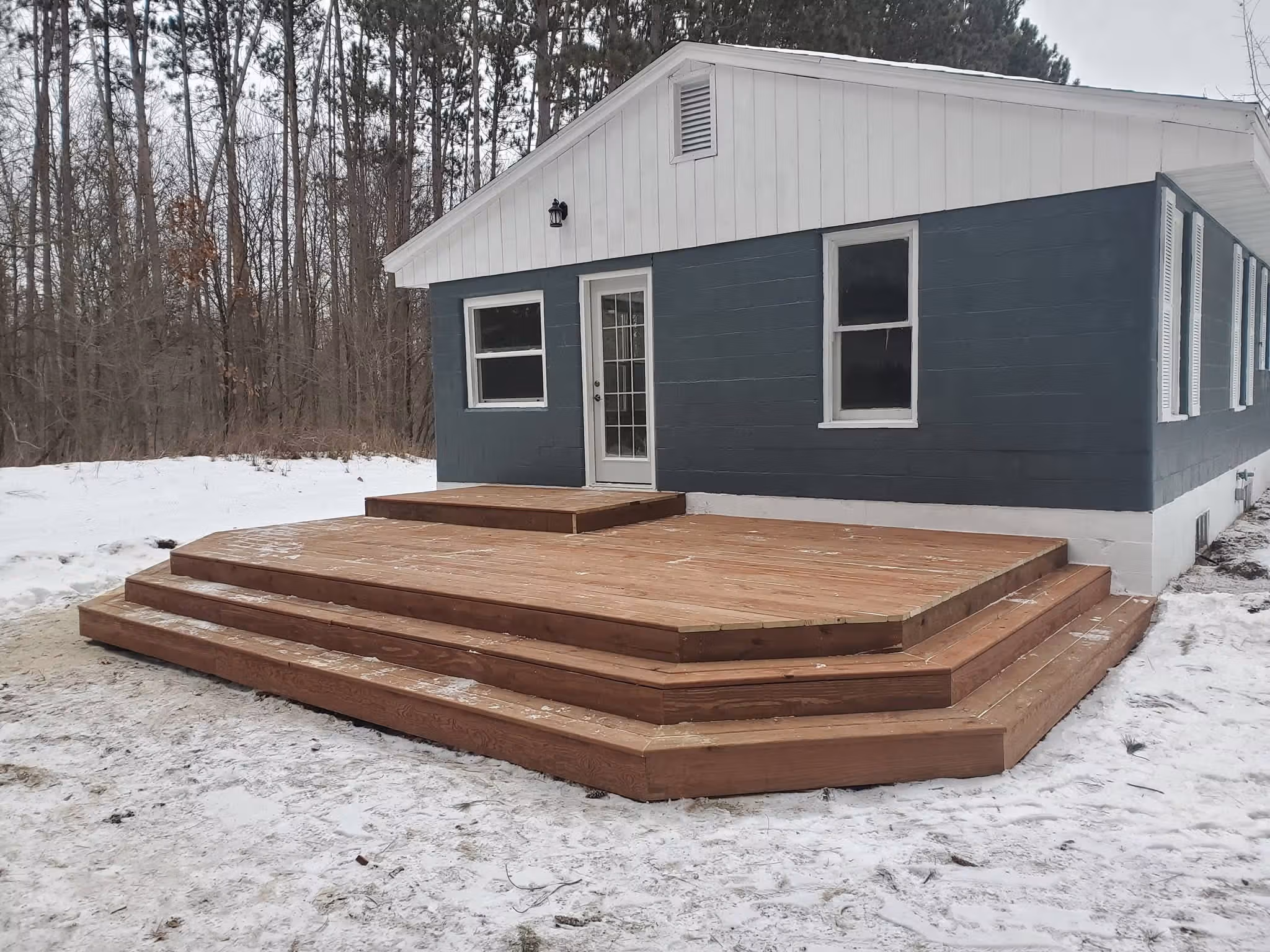 Large wooden deck with multiple steps in front of a blue and white house in a snowy yard.