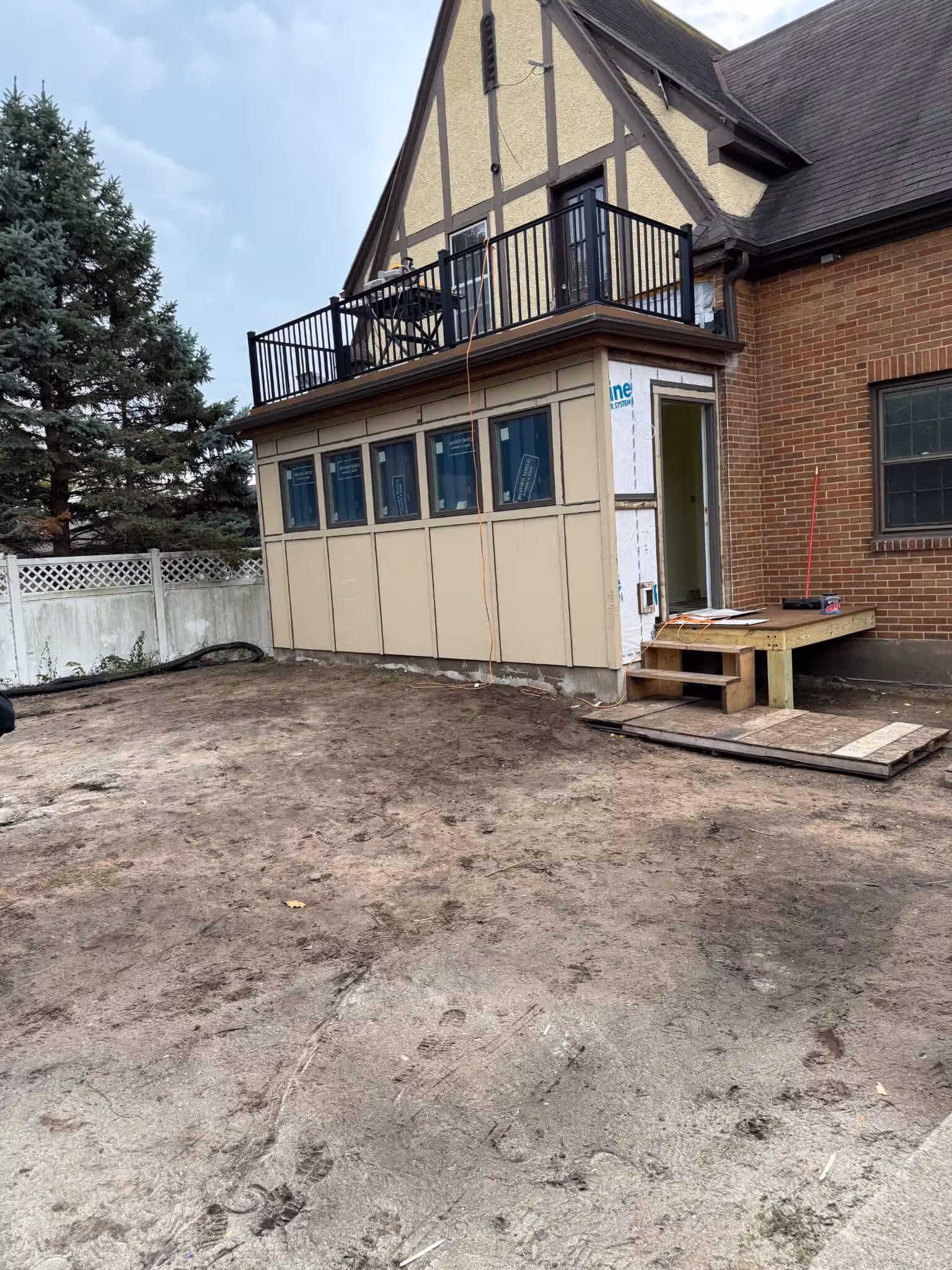 Backyard with dirt ground and a house extension featuring multiple windows and a balcony above.
