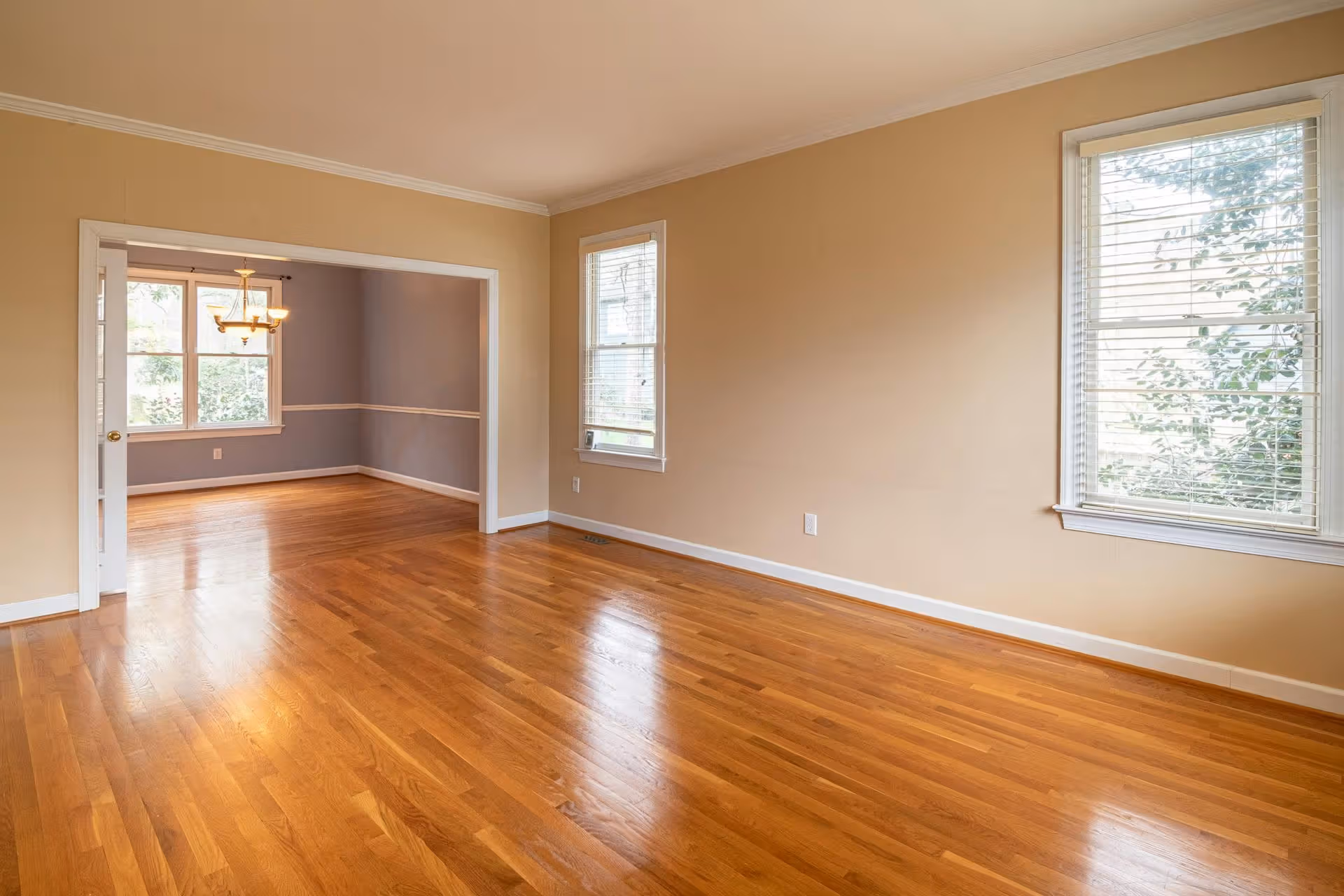 Empty room with polished hardwood floor, beige walls, and two windows with blinds.