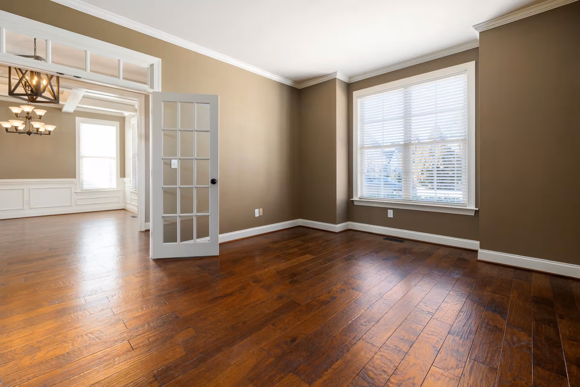 Empty room with brown walls, large window with white blinds, hardwood floors, and glass-paneled door.