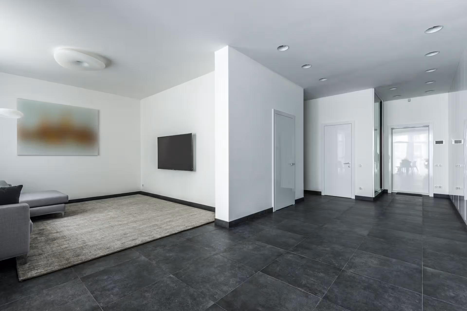 Minimalist room with gray tile floor, white walls, a gray couch, a blurred painting, and a wall-mounted TV.
