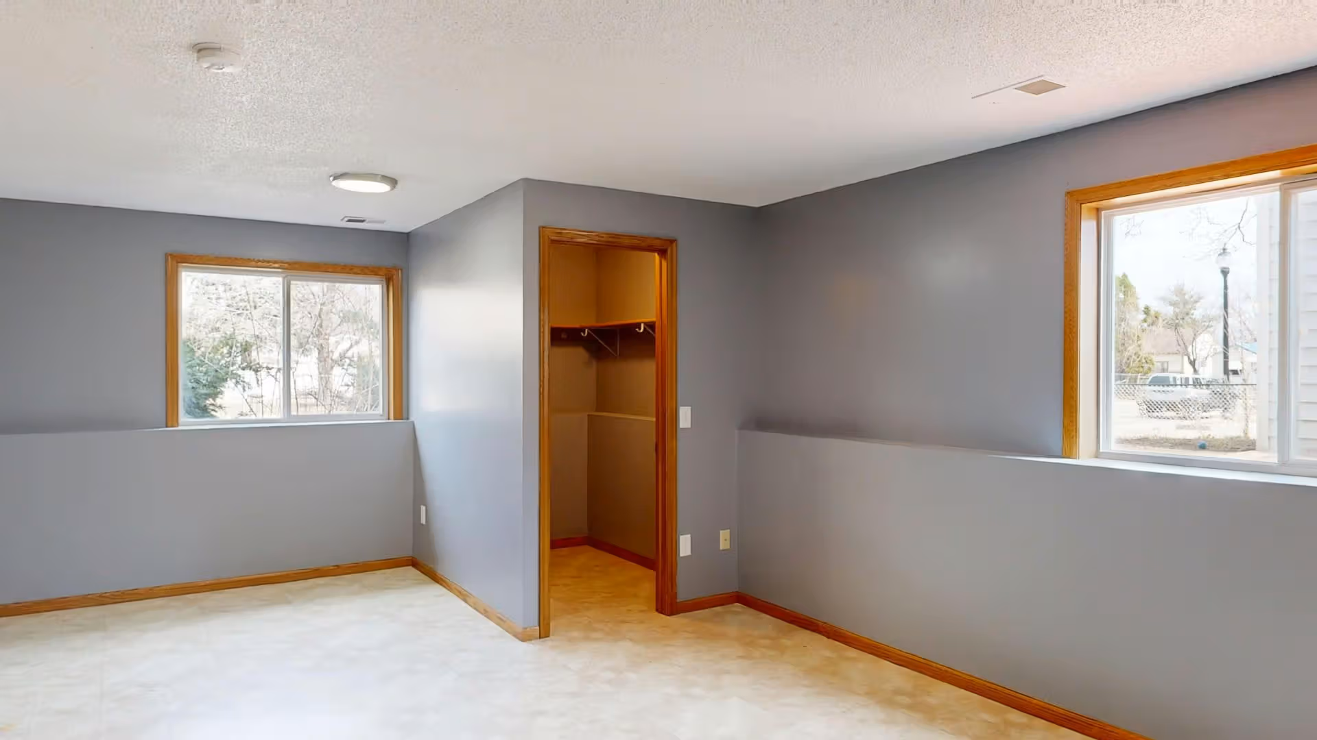 Empty room with gray walls, beige floor, two windows, and an open closet with wooden trim.