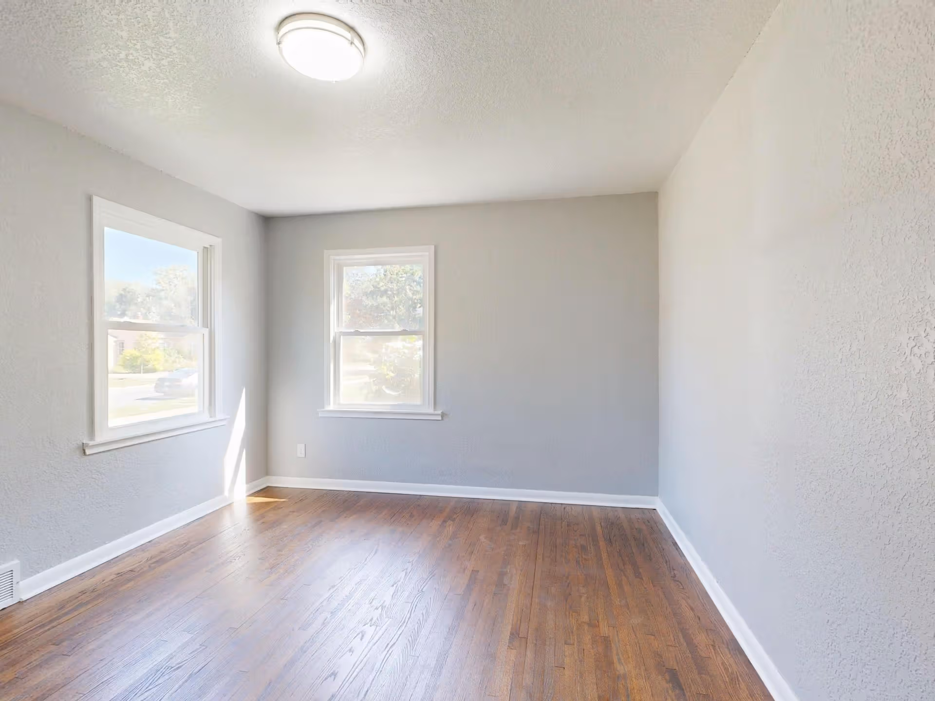 Empty room with two windows, gray walls, white trim, and hardwood flooring under ceiling light.