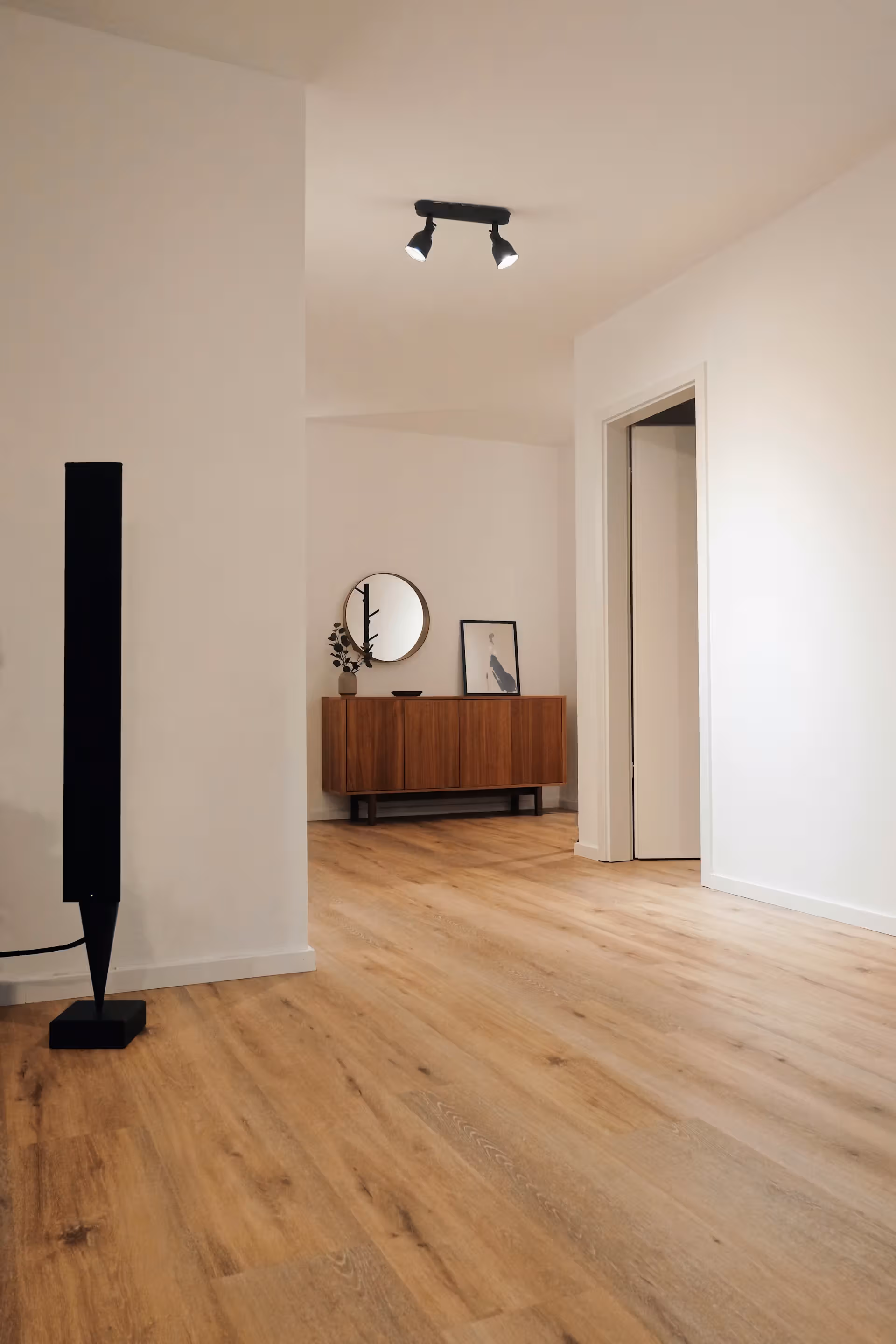 Room with light wood vinyl flooring, white walls, black floor lamp, wooden cabinet, round mirror, and framed art.